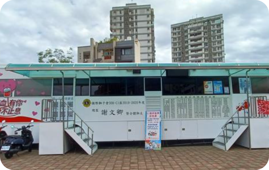 The image shows the 'Renhe Mobile Blood Donation Unit,' a fixed-point plug-in electric blood donation vehicle currently parked at Renhe Park in Beitun District, Taichung City, utilizing an external power source to reduce carbon emissions.
